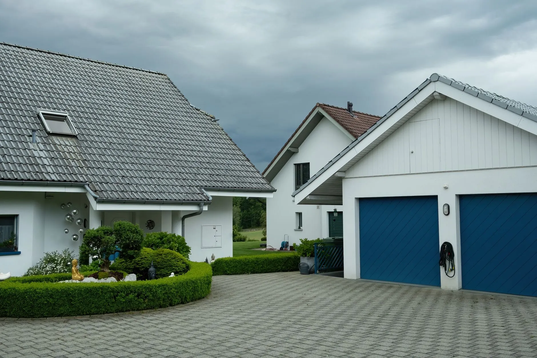 A white garage with two blue timber-clad doors beside a house with a clipped box hedge and a block-paved drive