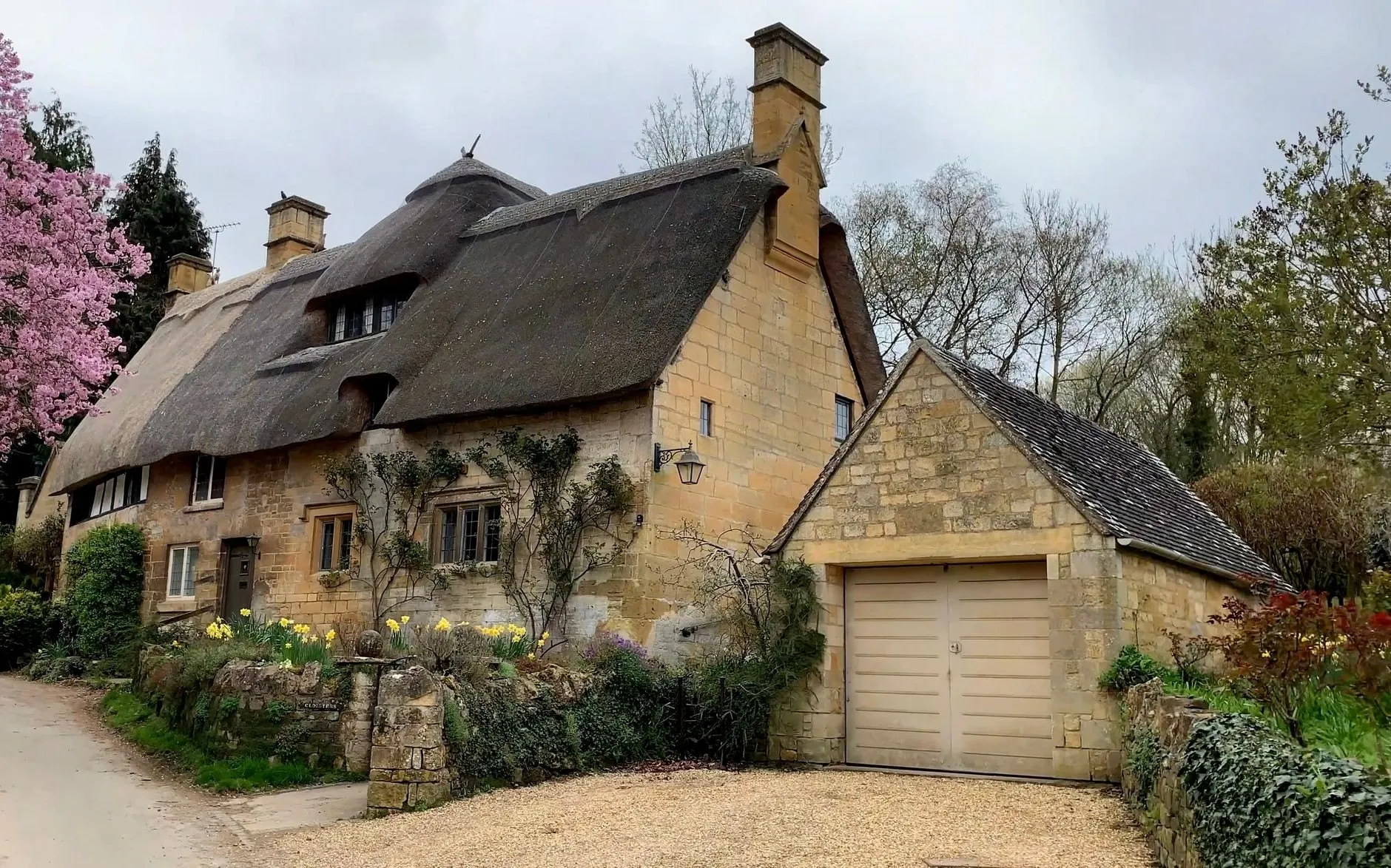 A thatched Cotswold stone cottage with daffodils by the path and a stone garage with a cream timber garage door