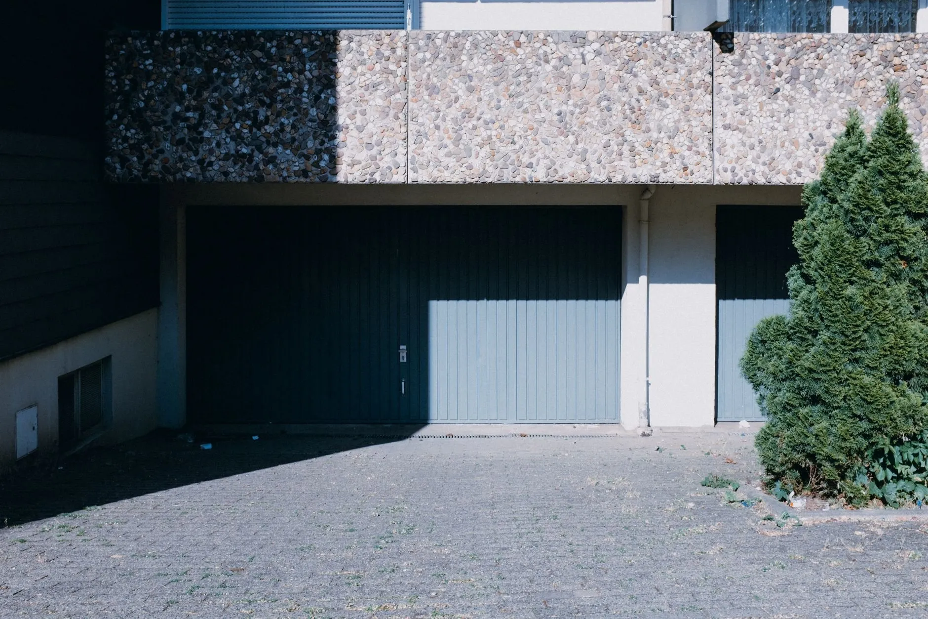 A deep blue-grey vertical-ribbed door under a pebble-dash overhang beside a conifer