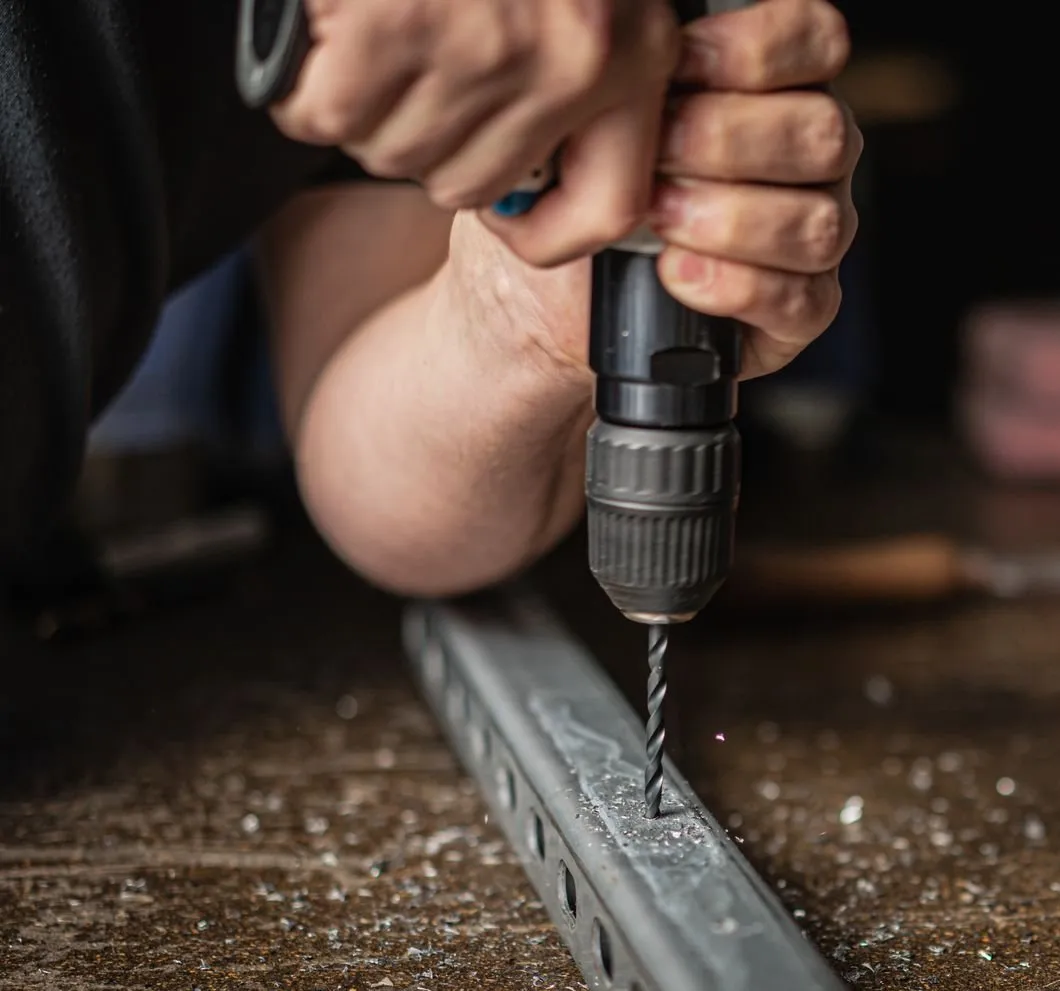 A hand drilling a fixing hole in a punched steel track on a workbench, with steel filings around it