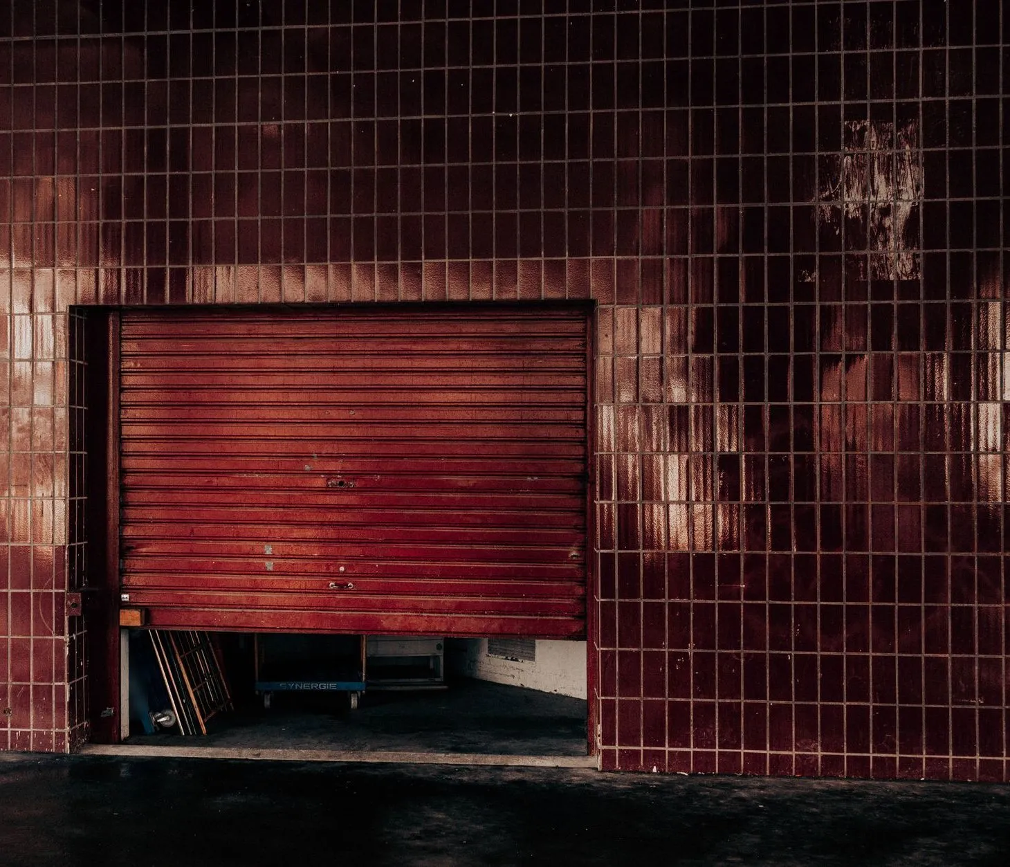 A red roller door stuck a foot above the ground in a tiled wall, with timber stacked inside