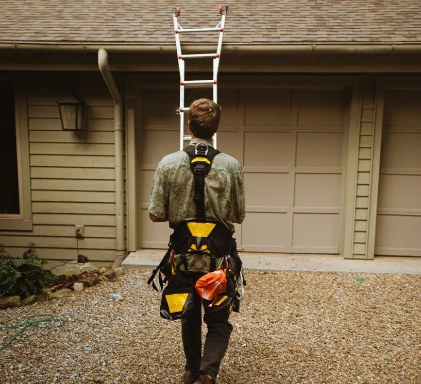 An engineer seen from behind, carrying his tool harness across a gravel drive towards a garage with a ladder up to the roof