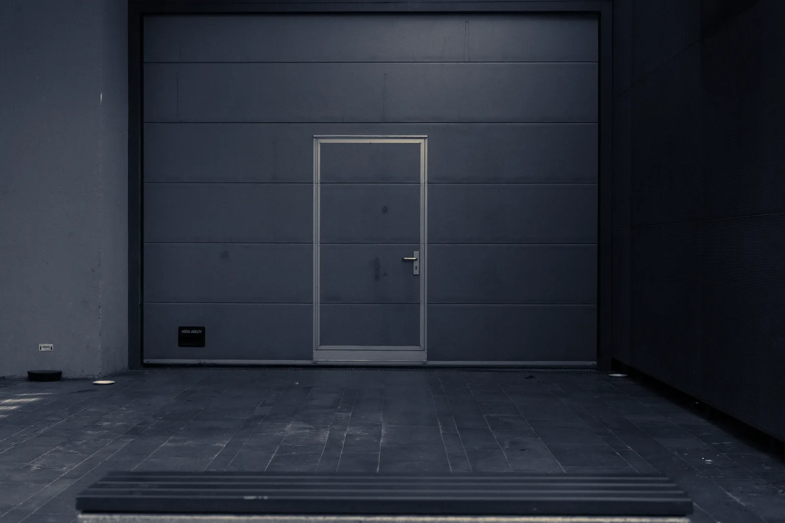 A sectional garage door closed in a concrete bay, cold steel shadow and a warm daylight edge.