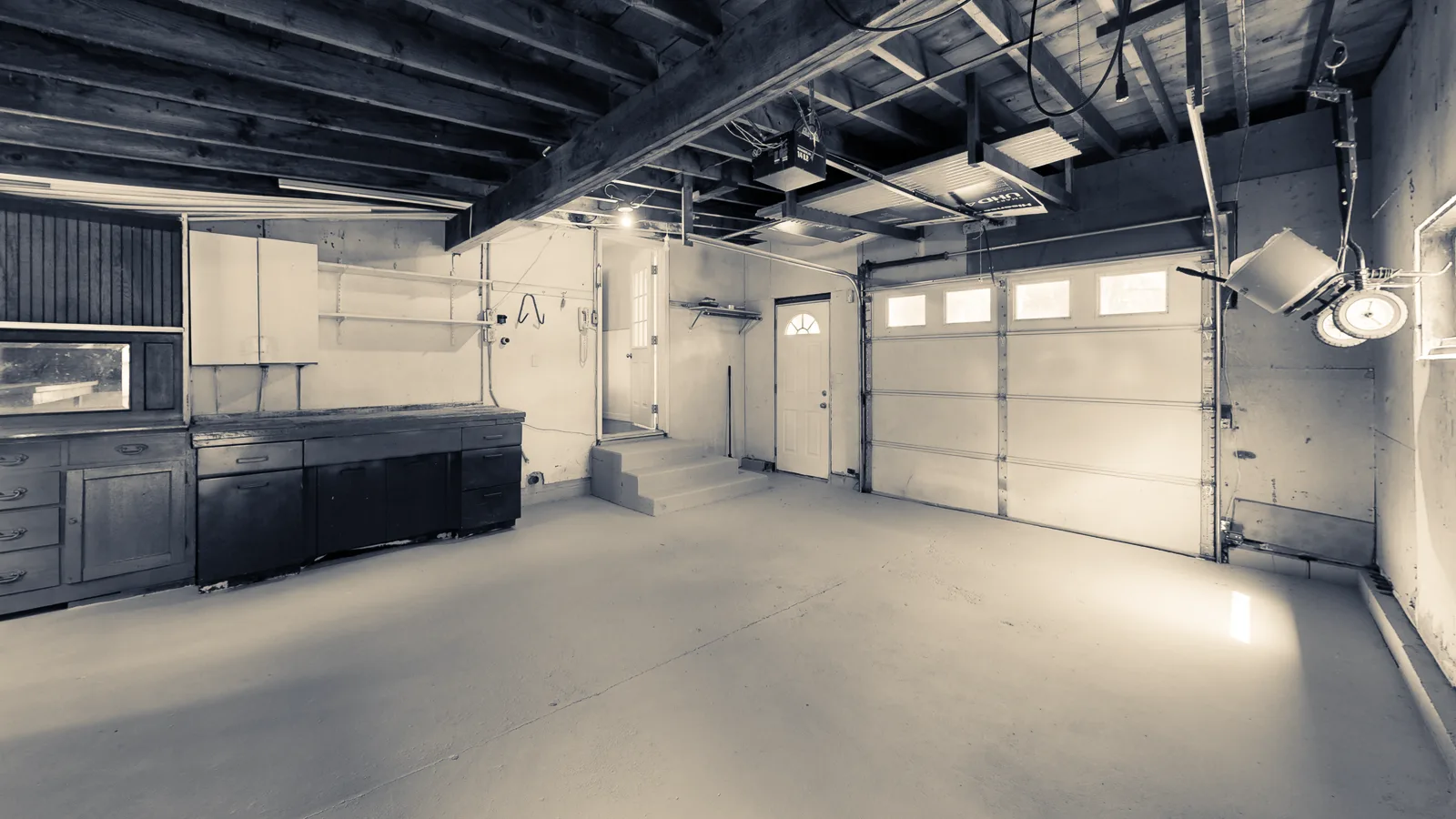 A garage interior showing a sectional door, its overhead track and the opener, cool concrete floor lit warm where daylight falls.